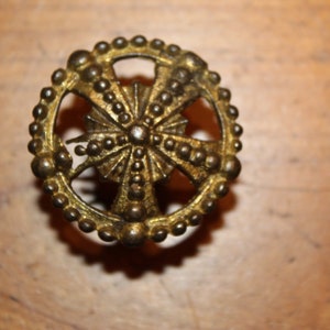 May include: Ornate, antique-style brass knob with a cross-shaped design at the center, surrounded by a ring of small, raised beads. The knob has a vintage aesthetic, suggesting it could be used for furniture or decorative purposes.