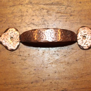 May include: A copper-colored cabinet pull with a textured, curved handle and decorative end pieces. The handle has an embossed pattern, and the end pieces have a bird-like shape with a hole for mounting. This hardware is suitable for furniture.