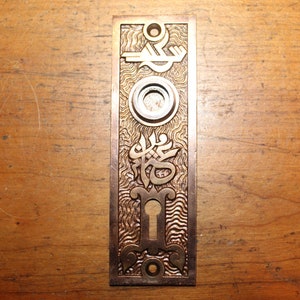 May include: A brass door plate with an ornate design. The plate has a keyhole and a round knob. The design includes a stylized key and a decorative pattern.