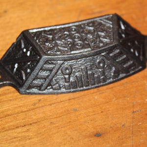 May include: A black, cast iron drawer pull with an ornate, geometric design. The pull has a rectangular shape with rounded ends and two circular holes for mounting. The surface is textured with detailed patterns.