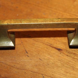May include: A brass cabinet pull with a rectangular shape and a slightly curved top. The pull has a brushed finish.