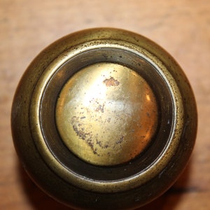 May include: Close-up of a vintage brass knob with a circular design. The knob has a rounded top and a recessed center, showing signs of age and wear. The brass has a warm, golden hue with darker areas.