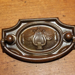 May include: A bronze drawer pull with an ornate design featuring a lyre, a musical instrument, in the center. The pull has a rounded shape and two small knobs on either side.