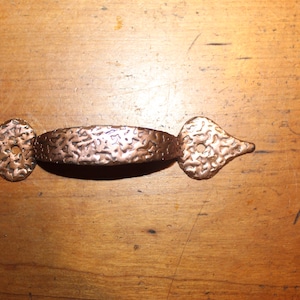 May include: A copper-toned metal drawer pull featuring a textured, hammered surface. The handle has a curved center grip and two flat, pointed ends, each containing a single circular screw hole for mounting to a cabinet or furniture piece.