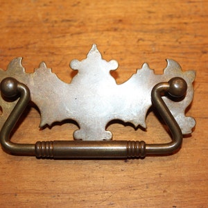 May include: A brass drawer pull with a decorative, ornate design. The pull has a curved handle and a decorative backplate with a stylized, floral pattern.