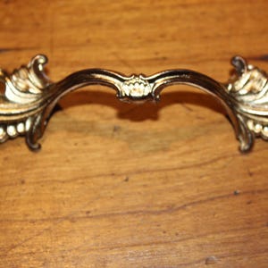 May include: Ornate gold-toned drawer pull with a curved handle and decorative floral accents. The hardware features a detailed design with a central flourish and symmetrical ends, suitable for furniture restoration.