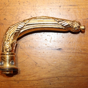 May include: A gold-colored, curved furniture handle with ornate detailing. The handle features a decorative, leaf-like design along its curved length and a small, rounded finial at the end. The base has a circular design with two visible screws.