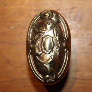 May include: Oval-shaped antique brass door knob with an ornate floral design. The knob features a detailed relief pattern with a central shield shape and swirling embellishments. The brass has a warm, aged patina.