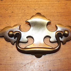 May include: Antique brass drawer pull with a decorative design. The pull features a central handle and ornate side elements. The hardware is set against a wooden surface.