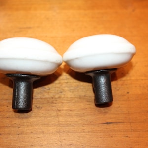 May include: Two white ceramic door knobs with black metal stems. The knobs are round and have a slight curve to them.