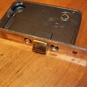 May include: A vintage metal door lock with a brass-colored faceplate and a dark, rectangular body. The lock features a sliding bolt and a keyhole. The object is set on a wooden surface.