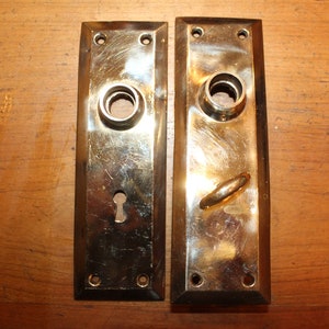 May include: Two antique brass door plates with keyholes and round holes for door knobs. The plates have a rectangular shape with rounded corners and are slightly tarnished.