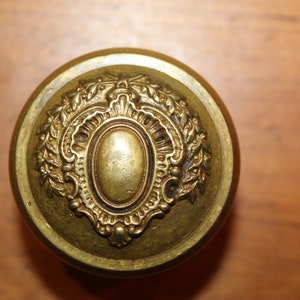 May include: A gold-toned door knob with an ornate design featuring a central oval and floral accents.