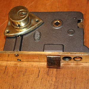 May include: A vintage door lock with a brass finish and a dark metal body. The lock features a keyhole, a latch, and a separate metal piece. The lock is likely for a door or cabinet.