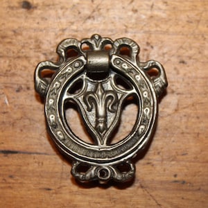May include: Ornate antique bronze drawer pull with a fleur-de-lis design. The circular pull features decorative scrollwork and a central emblem. The hardware is set against a wooden surface.