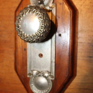 May include: Antique silver-toned door knob with a decorative rosette design, mounted on a dark brown, octagonal wooden backplate. The knob has a detailed, textured surface. A rectangular escutcheon plate is below the knob.