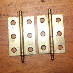 May include: Two brass hinges with four screw holes each, laid on a wooden surface. The hinges are rectangular with a central pin connecting the two plates. The brass has a slightly aged appearance.