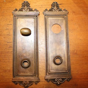May include: Two antique bronze door plates. One plate has an oval door knob, the other has a hole for a door handle. Both plates have a rectangular design with decorative edges and ornate top and bottom details.