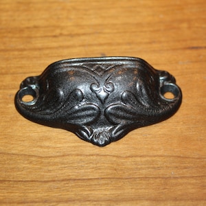 May include: Black metal drawer pull with an ornate design. The pull has two holes for mounting.