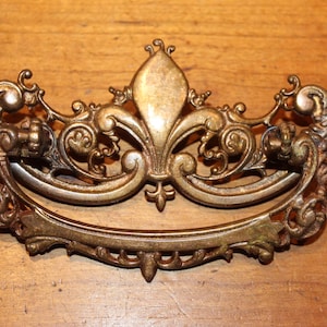 May include: Ornate antique brass drawer pull with a fleur-de-lis design. The handle features intricate scrollwork and a curved, decorative bar. The hardware is a warm, aged brass color, suitable for vintage furniture restoration.