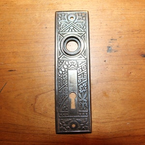 May include: An antique, rectangular door plate with intricate, embossed patterns. The metal plate features a keyhole, a circular opening, and decorative floral and geometric designs. The plate is a dark, aged metal, likely bronze or iron.