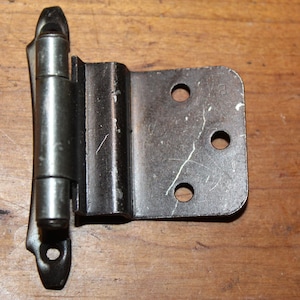 May include: A metal cabinet hinge with a rectangular plate and three screw holes. The hinge has a cylindrical barrel and a curved end piece. The metal has a dark, aged appearance, suggesting it is vintage or used.