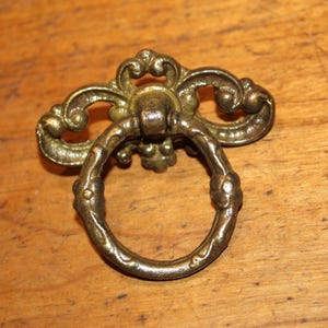 May include: Ornate brass drawer pull with a ring handle. The decorative hardware features a detailed floral design and is likely vintage. The pull is set against a wooden surface.