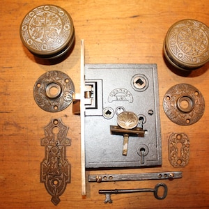 May include: Antique bronze door hardware set, including two doorknobs, a lock, key, and decorative plates. The doorknobs and plates feature an embossed floral design. The lock has the text "SARGENT" and "A.90".