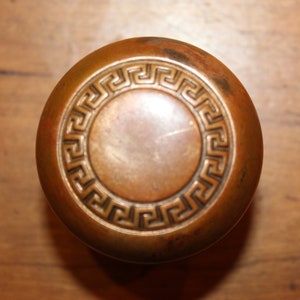 May include: A round, brass door knob with a decorative, raised, Greek key pattern around the edge.