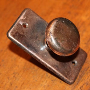 May include: Close-up of an antique door knob with a rectangular backplate and a rounded knob. The metal hardware has a tarnished, aged appearance, with a warm, bronze-like color. The knob is mounted on a wooden surface.