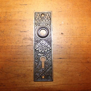 May include: An antique, rectangular door plate with intricate, embossed floral and geometric patterns. The metal plate has a keyhole, a circular knob opening, and two screw holes. The color is a muted bronze.