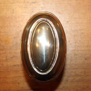 May include: Oval-shaped brass cabinet knob with a polished finish. The knob has a raised central oval and a decorative outer rim. The hardware is designed for use on drawers or cabinet doors.