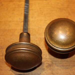 May include: Two antique brass door knobs. One knob is attached to a metal rod. The other knob is a separate, round piece. Both knobs have a warm, aged patina and are set against a wooden surface.