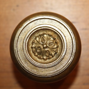 May include: A brass door knob with a decorative floral design in the center. The knob has a circular shape with concentric rings around the center design.