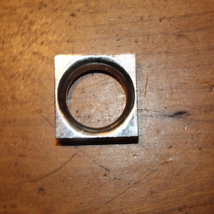 May include: A square metal object with a circular hole in the center. The object is silver in color and appears to be made of a solid metal. The object is sitting on a wooden surface.