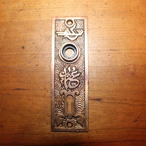 May include: A brass door plate with an ornate design featuring a keyhole, a circle, and a decorative pattern. The plate has a dark brown patina.