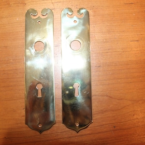 May include: Two vintage brass door plates. Each plate is rectangular with a keyhole, a round hole, and a decorative top and bottom. The brass has a warm, golden hue and shows signs of age.