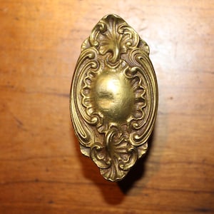 May include: Ornate brass door knob with an oval shape and detailed scrollwork. The knob has a central, sunburst-like design and is set against a wooden surface. The antique style suggests a vintage or classic aesthetic.