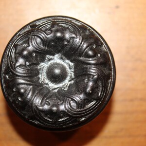 May include: A black, ornate cabinet knob with a detailed floral and scroll design. The knob has a central, raised circular element and a decorative, textured surface. The hardware is likely made of metal.