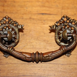 May include: Antique bronze drawer pull with a decorative sunburst design. The pull features a curved handle with detailed leaf and scrollwork. The decorative elements include a sunburst design and ribbon accents. This hardware is suitable for cabinets and furniture.