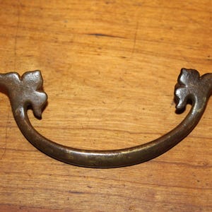 May include: A vintage, ornate, brass drawer pull with a curved shape and leaf-shaped ends.