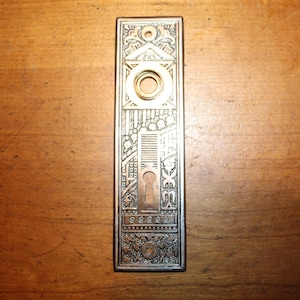 May include: An antique rectangular door plate with an ornate design. The plate is made of metal with a bronze finish and features a keyhole, a circular opening, and detailed engravings of architectural and floral elements.