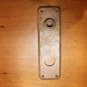 May include: A brass door plate with two round holes, one larger than the other. The plate has a slightly rounded rectangular shape.