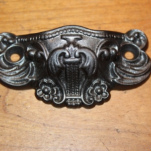 May include: Black cast iron drawer pull with ornate floral design. The pull has two holes for mounting.