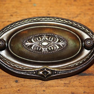 May include: An oval-shaped, antique-style cabinet pull with a decorative floral design in the center. The pull is made of metal and has a dark bronze finish.