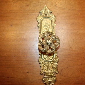 May include: A gold-toned door handle with an ornate design. The handle is a single knob with a floral pattern and is attached to a decorative backplate.