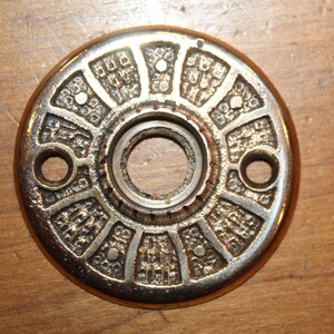 May include: A round, brass door knob escutcheon with a decorative, geometric design. The escutcheon has a central hole for the knob.