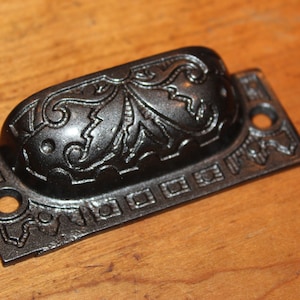 May include: Black cast iron drawer pull with an ornate design. The pull has a rectangular base with a rounded top and a decorative border.