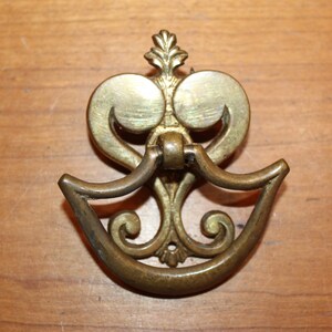 May include: Antique brass drawer pull with a decorative design. The pull features a curved handle and ornate detailing, including a floral element at the top. The hardware is mounted on a wooden surface.