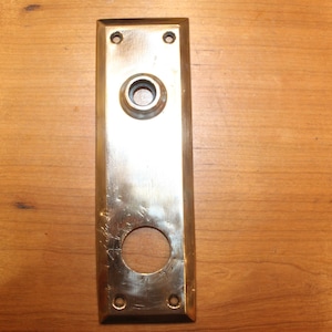 May include: A brass door plate with a round hole for a doorknob and a rectangular hole for a keyhole.
